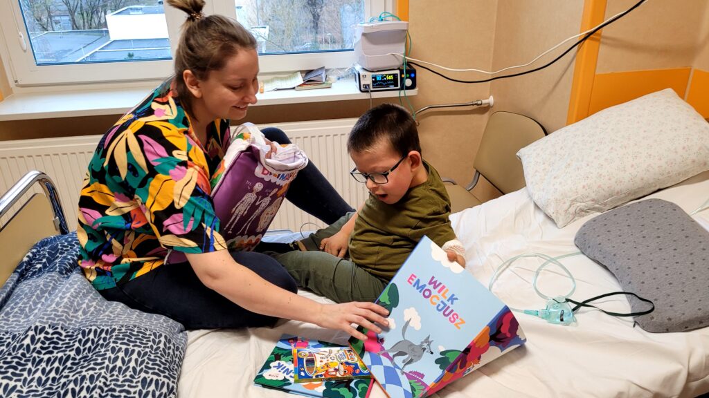 Chłopczyk z mamą na szpitalnym łóżku trzymający książeczki podarowane przez Fundację KIDS